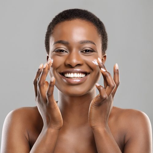 Skin Care Concept. Portrait Of Happy Black Woman Applying Moisturizing Cream on Face, Grey Studio Background