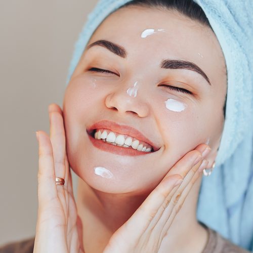Happy smiling pretty beautiful woman girl with towel on head smiling touch healthy clean soft hydrated skin care after applying cream on young face natural beauty. without make up long banner